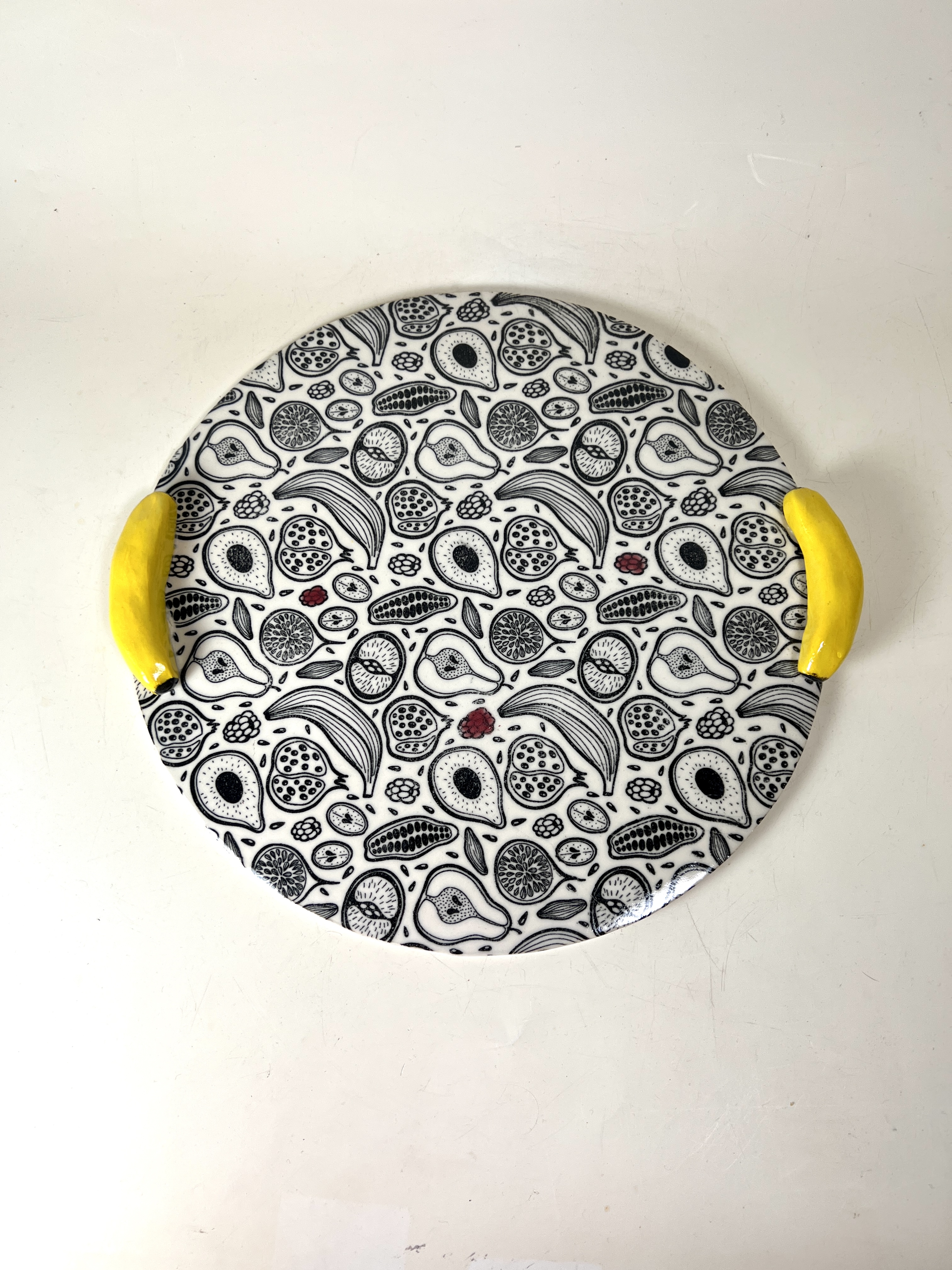 Round ceramic cake plate with dense black and white fruit transfer print and yellow handles